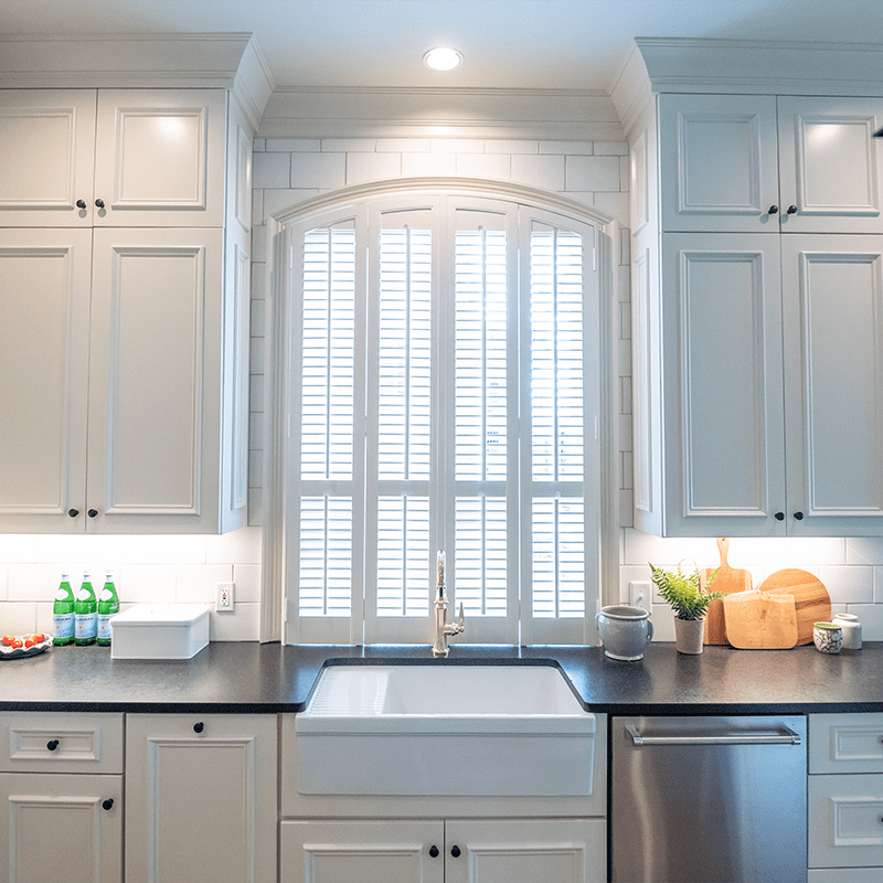 White interior shutter sets built by Dwell Shutter & Blinds displayed in a beautiful kitchen with white cabinets and backsplash tile.