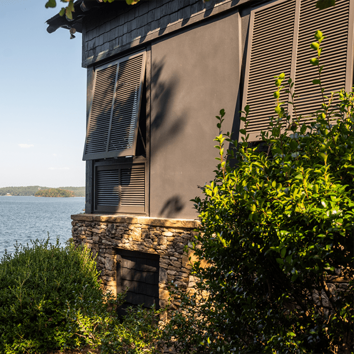 Brown Bahama exterior shutters built by Dwell Shutter & Blinds displayed on a lake house.